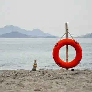 Salvagente rosso di salvataggio posato sulla sabbia, vicino a uno specchio d’acqua che può essere mare, lago o fiume.