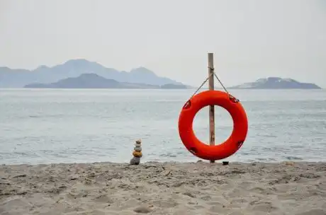 Salvagente rosso di salvataggio posato sulla sabbia, vicino a uno specchio d’acqua che può essere mare, lago o fiume.